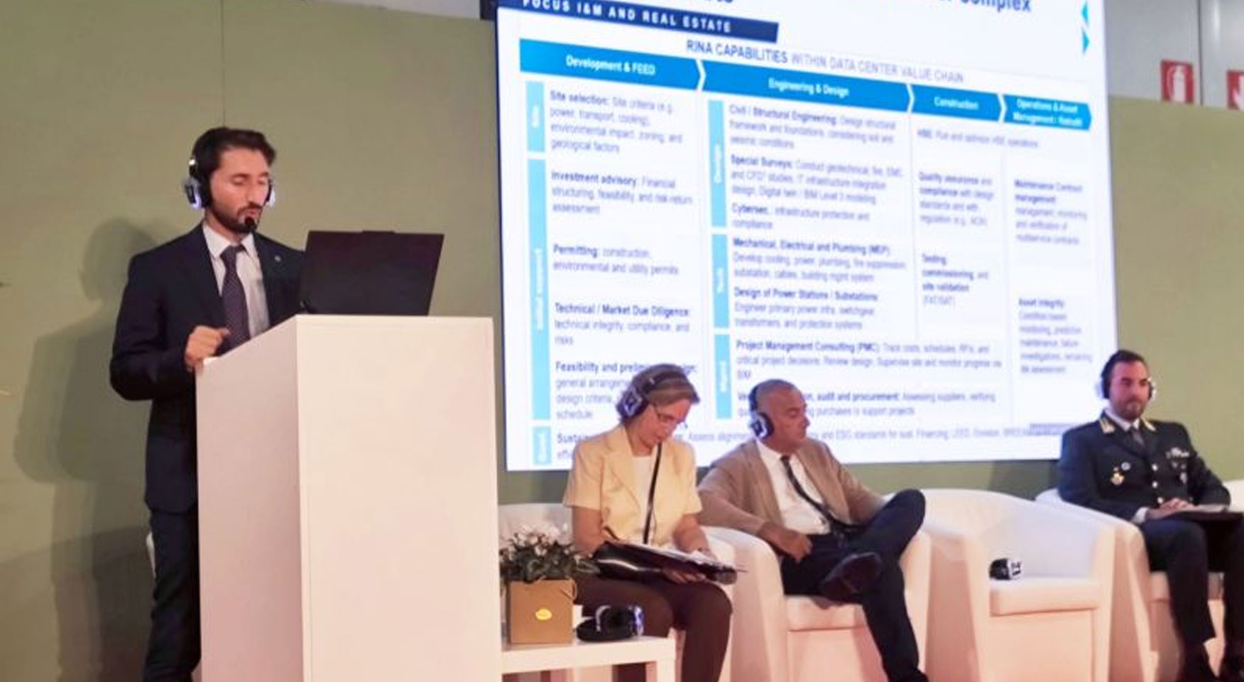 Relatore durante una conferenza sulle infrastrutture digitali con panel di partecipanti e schermo di presentazione.