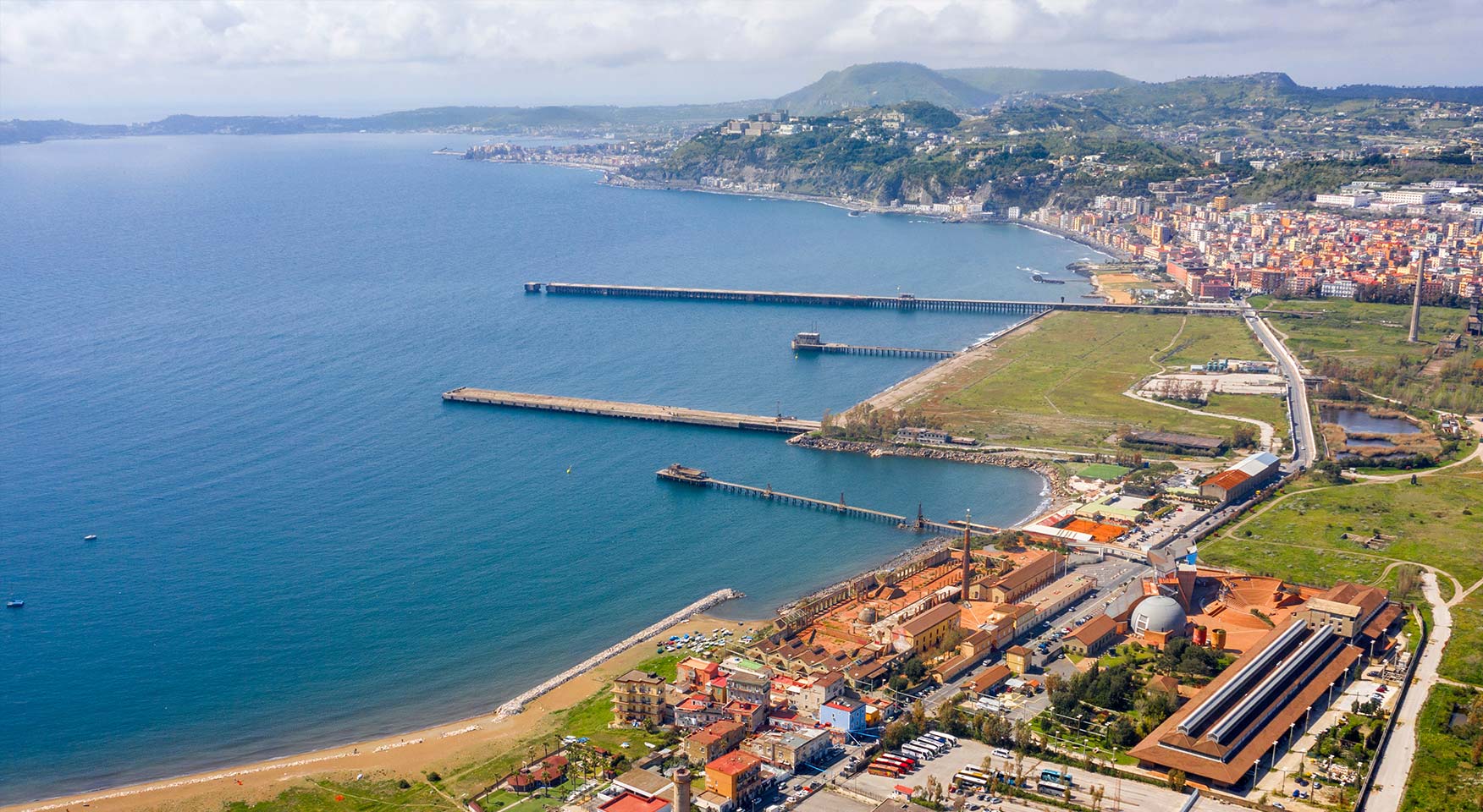 Vista aerea di un waterfront costiero con moli, litorale, area urbana e colline affacciate sul golfo.