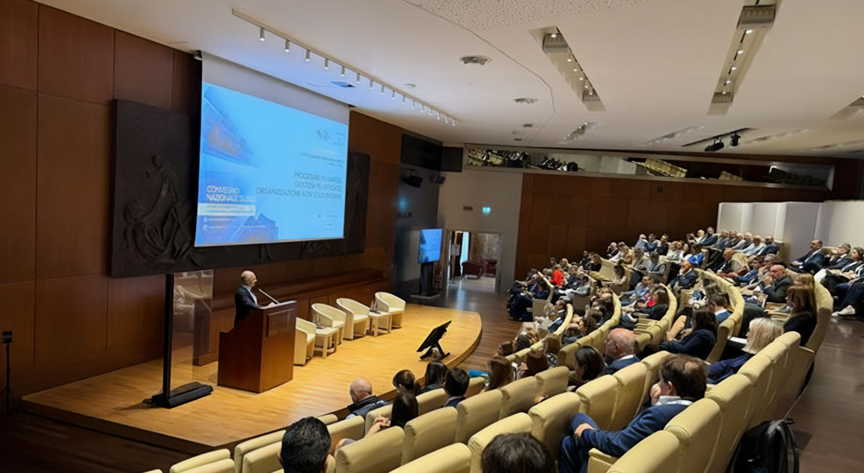 Relatore durante una conferenza in auditorium con numerosi partecipanti seduti in sala.